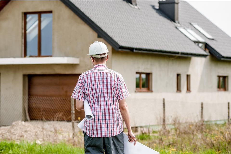 Fachmann mit Bauplänen und Schutzhelm vor einem Einfamilienhaus